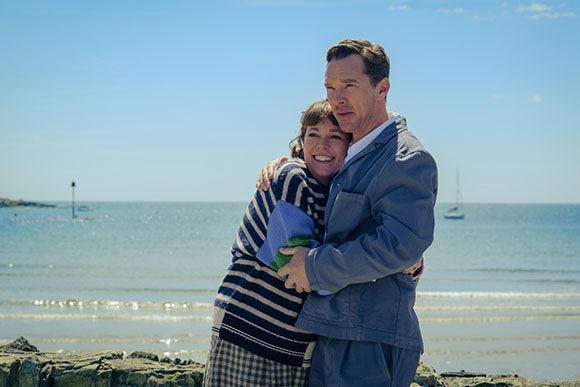 A man in a light blue jacket hugs a woman in a striped sweater on a beach by the ocean.