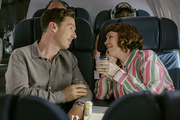 A man in a grey shirt and a woman in a pink striped shirt sit in airplane seats, talking and holding drinks.