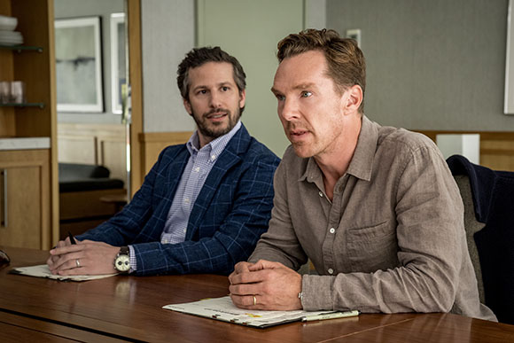 Two men sit at a wooden table in an office, looking toward something off-camera.