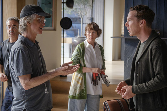 A man in a grey polo gestures while talking to a man in a dark cardigan and a woman in a green patterned cardigan.
