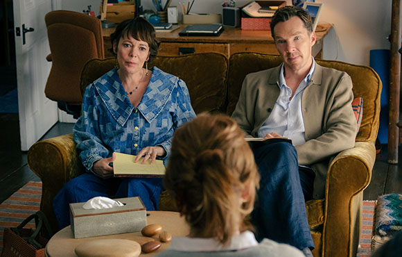 A woman in a blue denim shirt and a man in a tan blazer sit on a brown couch facing a person with blonde hair.