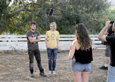 A film crew uses a boom mic and camera to film two people standing in a dirt field near trees.