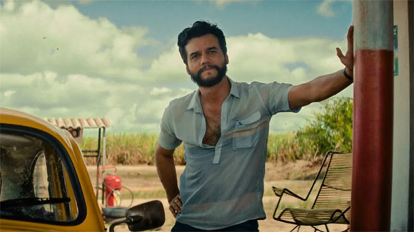 A bearded man in a light blue short-sleeved shirt leans against a red and white post near a yellow car.