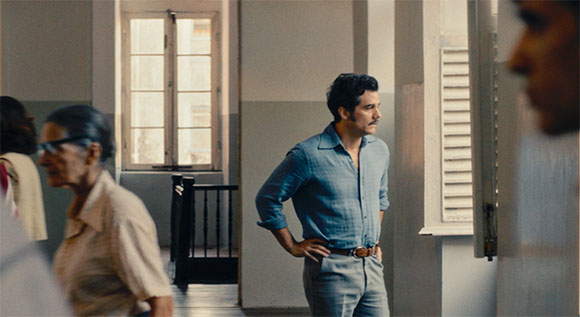 A man in a blue button-down shirt stands in a hallway, looking to the side.