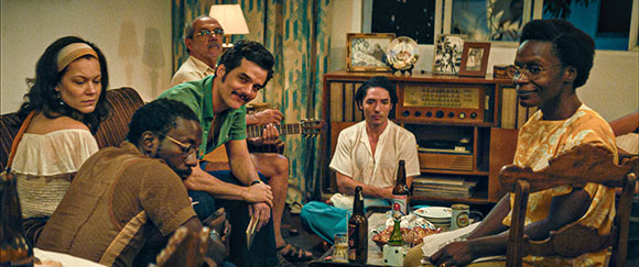 A diverse group of people sitting in a living room with a man playing a guitar.