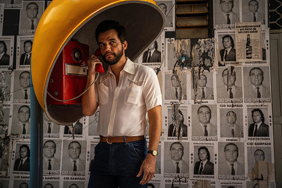 A man in a white short-sleeved shirt talks on a red rotary phone inside a yellow booth against a wall of posters.
