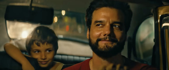 A man with a beard smiles while a young boy sits beside him in a car at night.