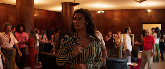 A woman in a striped shirt looks away from a crowd of people in a dimly lit, wood-paneled room.