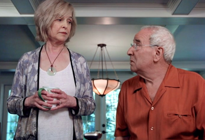 A blonde woman in a patterned cardigan holds a mug while looking at an older man in an orange shirt.
