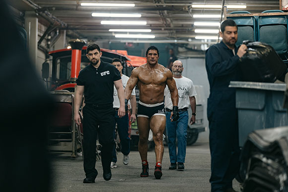 A muscular shirtless man walks through a garage flanked by men in black and white clothing.