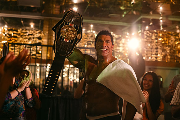 A shirtless, muscular man smiles while holding a large black and gold championship belt aloft.
