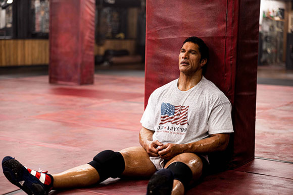 A man in a white t-shirt and knee pads sits on a red gym floor, leaning against a red padded pillar.