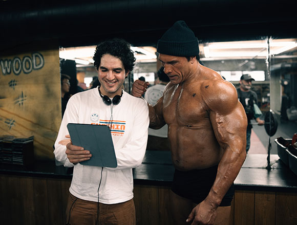 A man in a white shirt holds a tablet while showing it to a muscular man in a black beanie.