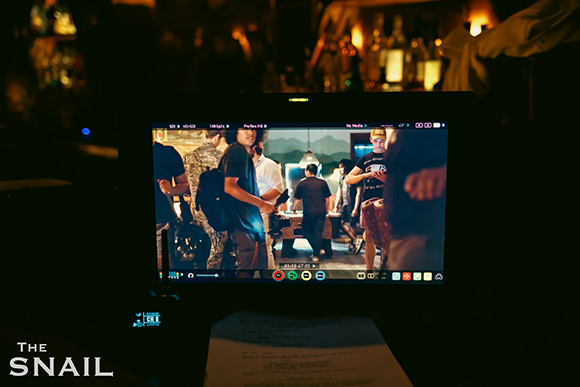 A film monitor shows a group of people in a dimly lit room, set against a dark background.