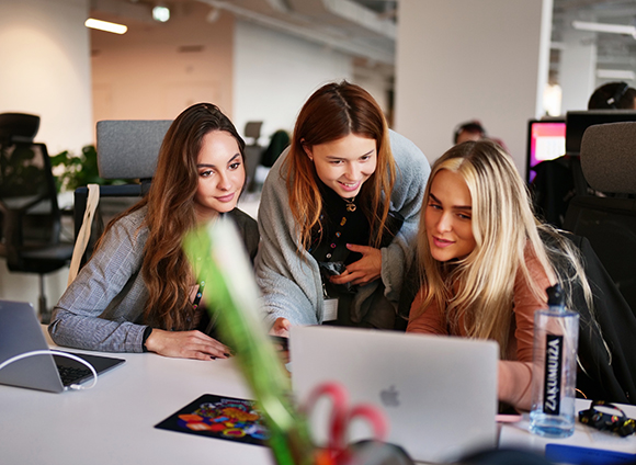 Three young women gather around a laptop in a bright, modern office space.
