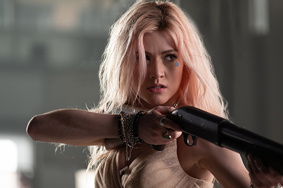 A woman with long pink hair holds a black shotgun with both hands.