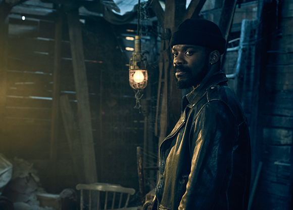A man with a beard wearing a dark beanie and leather jacket stands in a dimly lit room.