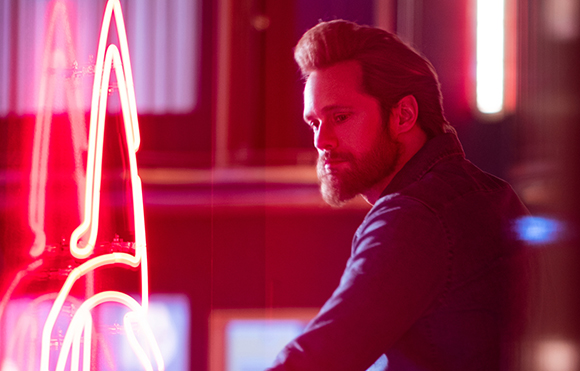 A bearded man in profile looking toward a bright red neon sign in a dimly lit setting.