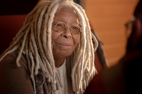 Elderly woman with long white dreadlocks and gold-rimmed glasses looking at the camera.