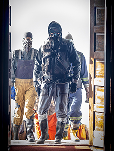 Three individuals in dark tactical gear and protective clothing walk through an open doorway.