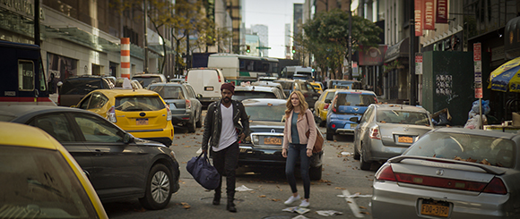 A man in a leather jacket and a woman in a pink jacket walk through a congested city street with many cars.