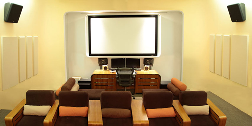 A media control room with a large projection screen, studio monitors, and rows of cushioned seating.
