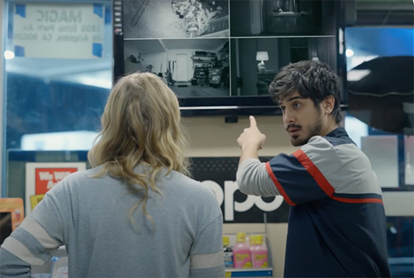 A man points toward a wall of security monitors while talking to a woman with blonde hair.
