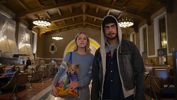 A woman holding a small dog and a man in a hoodie stand in a large, high-ceilinged hall.