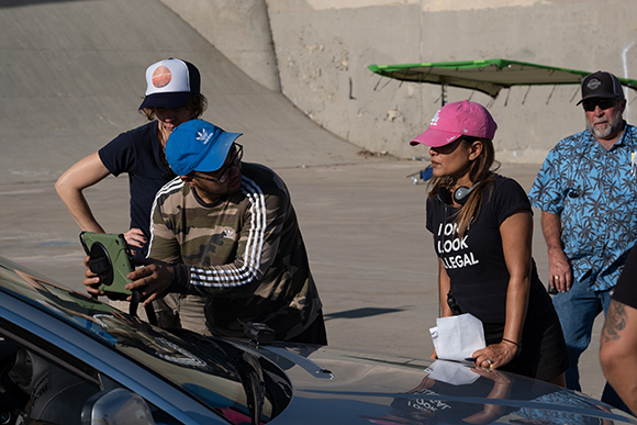A group of people stand around a car, looking at a tablet held by a person in a blue cap.