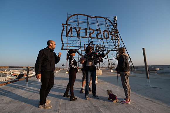 A film crew with a camera and tripod works on a rooftop in front of a large metal Brooklyn sign.