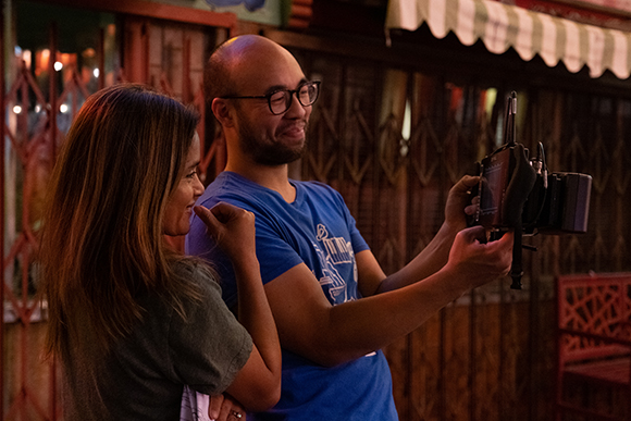 A man in a blue shirt and glasses holds a camera monitor while a woman looks on beside him.