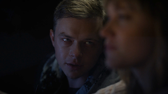 A man with short hair looks intensely toward a woman in the foreground in a dark setting.