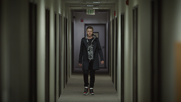 A man in a dark coat and patterned shirt walks toward the camera in a long, narrow hallway.