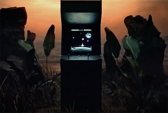 A lit arcade machine stands amidst large leaves and rocks under an orange sunset sky.