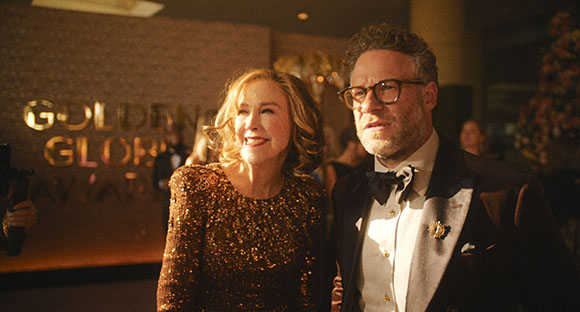 A man in a tuxedo and glasses stands next to a woman in a sparkling gold dress at an awards ceremony.