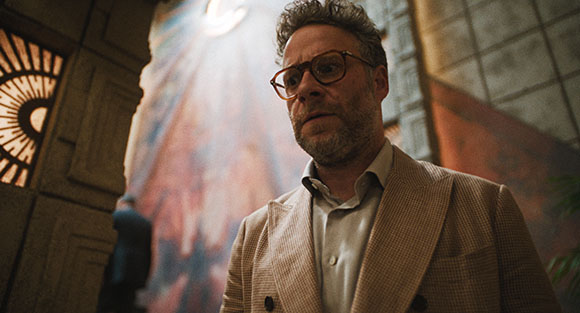 A man with glasses and a beard wearing a tan blazer looks thoughtfully to the side.