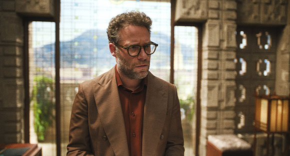 A man with curly hair and glasses wearing a brown blazer and reddish shirt stands indoors.