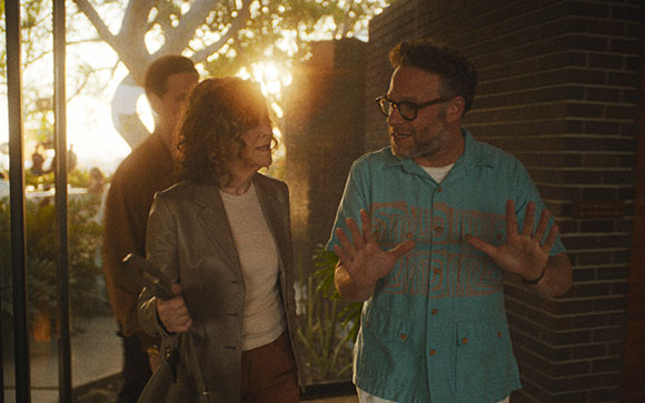 A man in a blue shirt gestures with both hands while talking to a woman in a grey blazer outdoors.