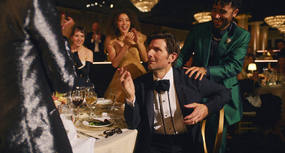 A man in a tuxedo gestures at a formal dinner table while guests clap and smile in the background.