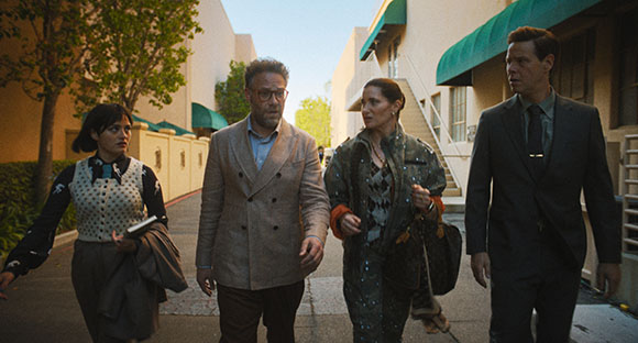 Four people walk together along a narrow outdoor path between light-colored buildings.