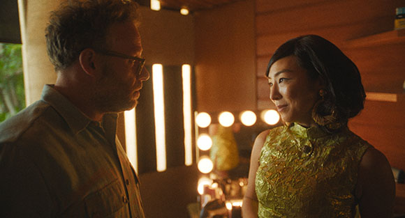 A man in a collared shirt and a woman in a yellow patterned dress look at each other in warm lighting.