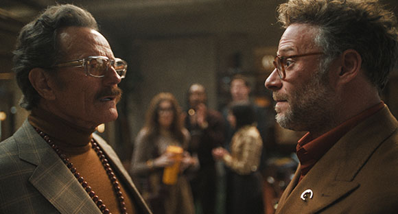 Two men with glasses and facial hair face each other in a crowded, dimly lit indoor setting.