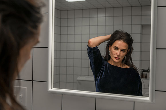 A woman with long dark hair looks at her reflection in a bathroom mirror while touching her hair.
