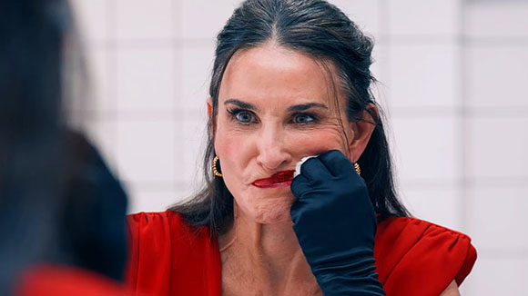 A woman in a red dress looks into a mirror while a gloved hand applies red lipstick to her mouth.