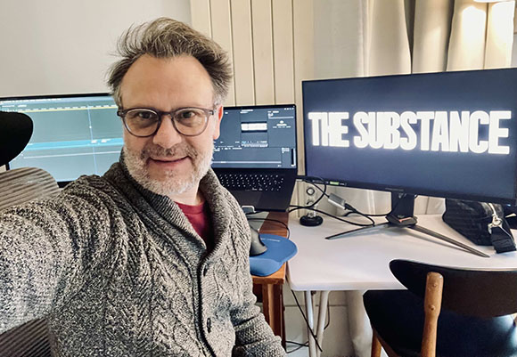A man in a grey cardigan takes a selfie in front of computer monitors in an office.