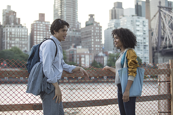 A young man with a backpack and a young woman with curly hair talk by a chain-link fence in a city.