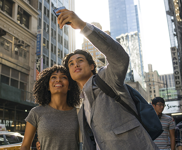 A young man in a grey blazer takes a selfie with a smiling woman in a grey t-shirt on a city street.