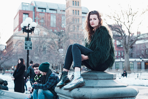 A woman with curly hair sits on a stone pedestal in a city park with buildings and trees in the background.