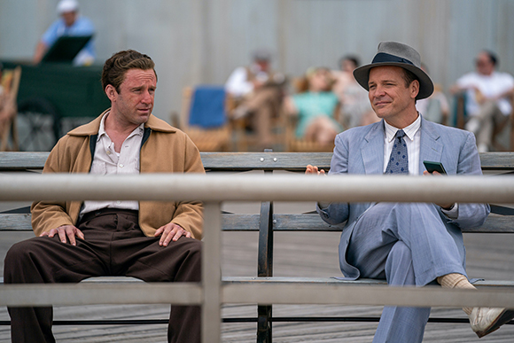 A man in a tan jacket and a man in a light blue suit and fedora sit on a wooden bench.