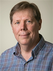 A man with short brown hair and blue eyes wearing a blue and white plaid button-down shirt.
